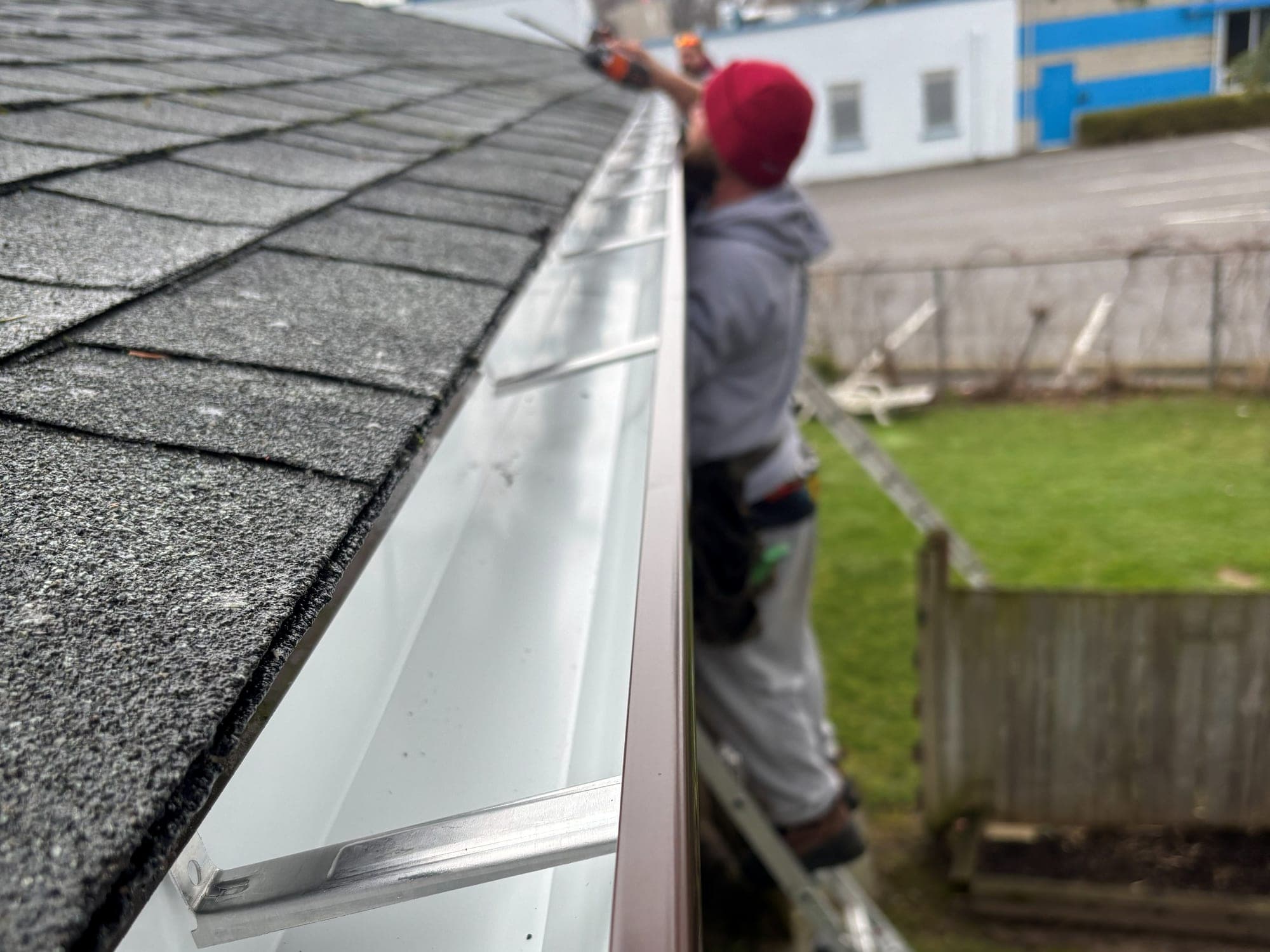 Crew member on a ladder fastening a newly hung eavestrough, brown outside and white inside with hidden hangers, along an asphalt shingle roof edge
