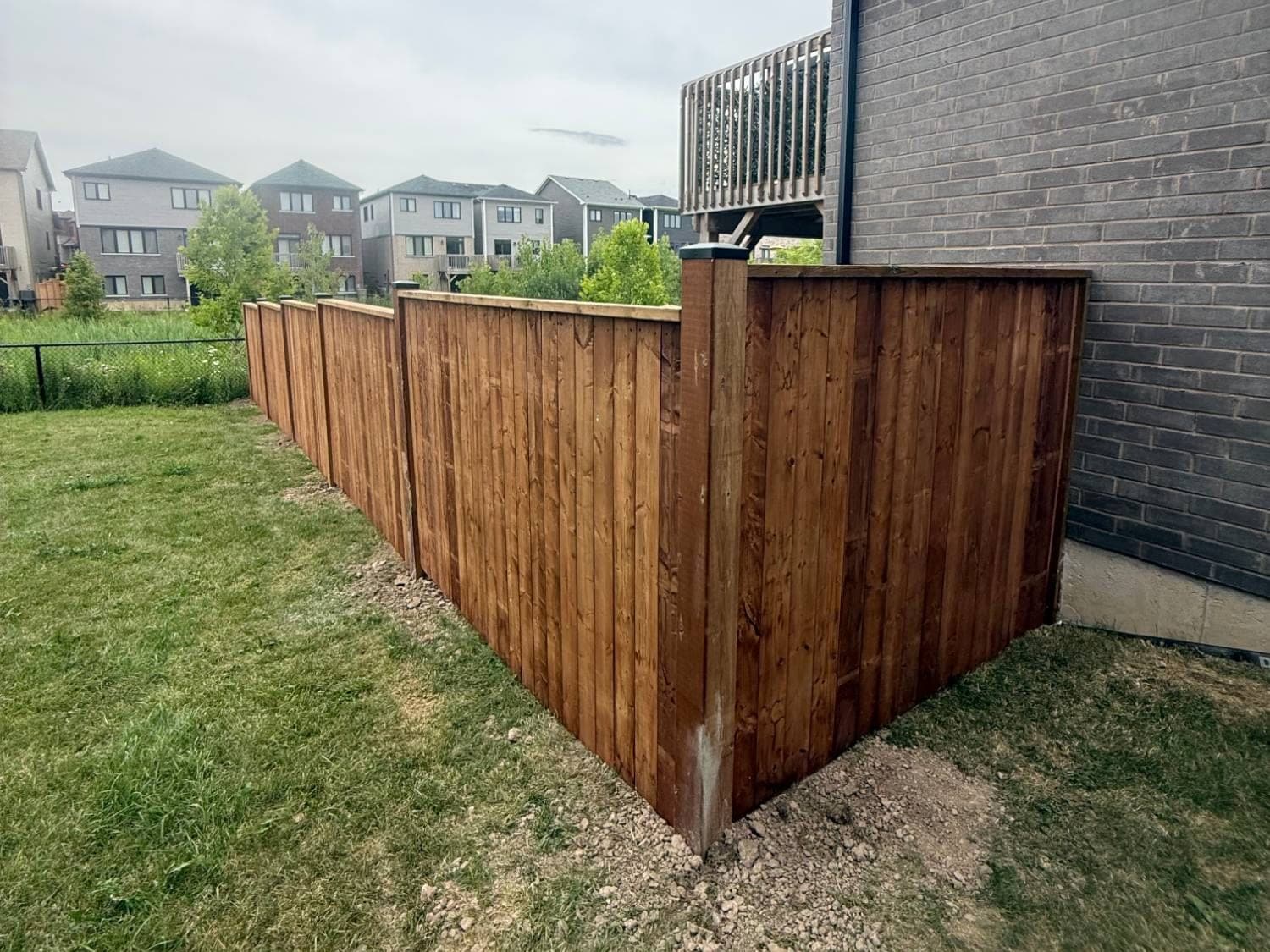New stained wood privacy fence with capped posts, running along a backyard and turning a corner to meet the brick wall of the house