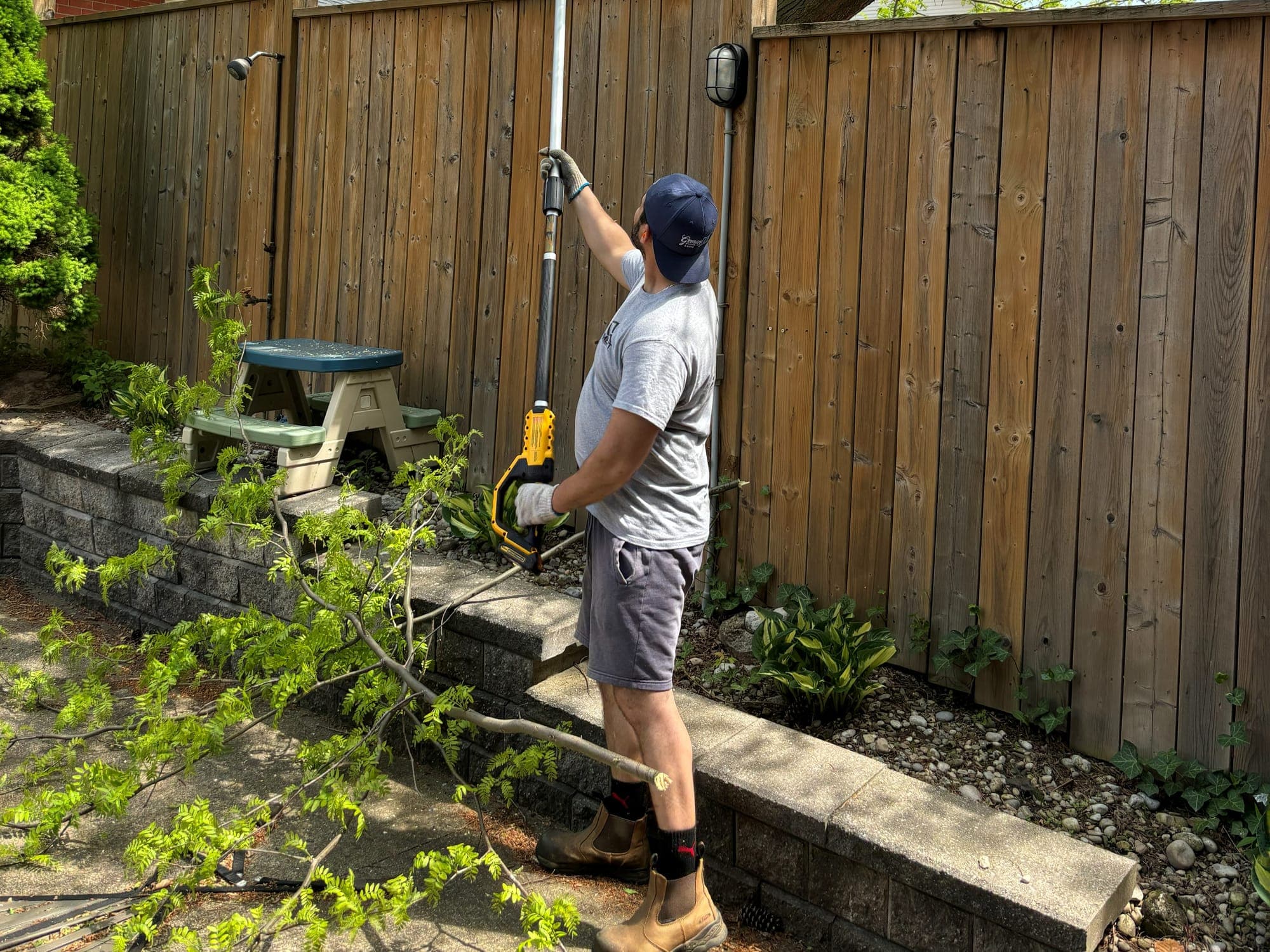 Crew member cutting back overhanging tree branches with a pole saw beside a backyard fence at a managed property in Niagara Falls