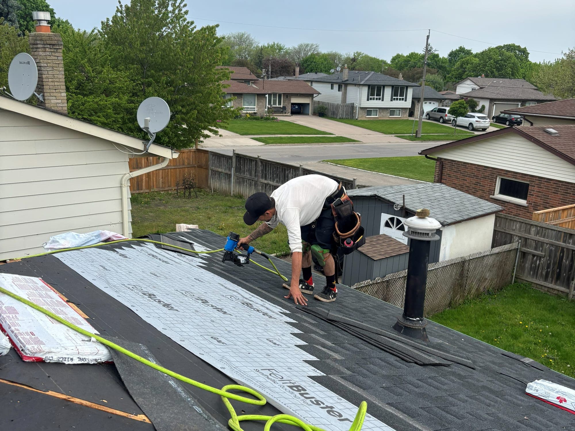 Roofer with a nail gun fastening new asphalt shingles over fresh synthetic underlayment on a residential roof, with the air hose coiled across the deck